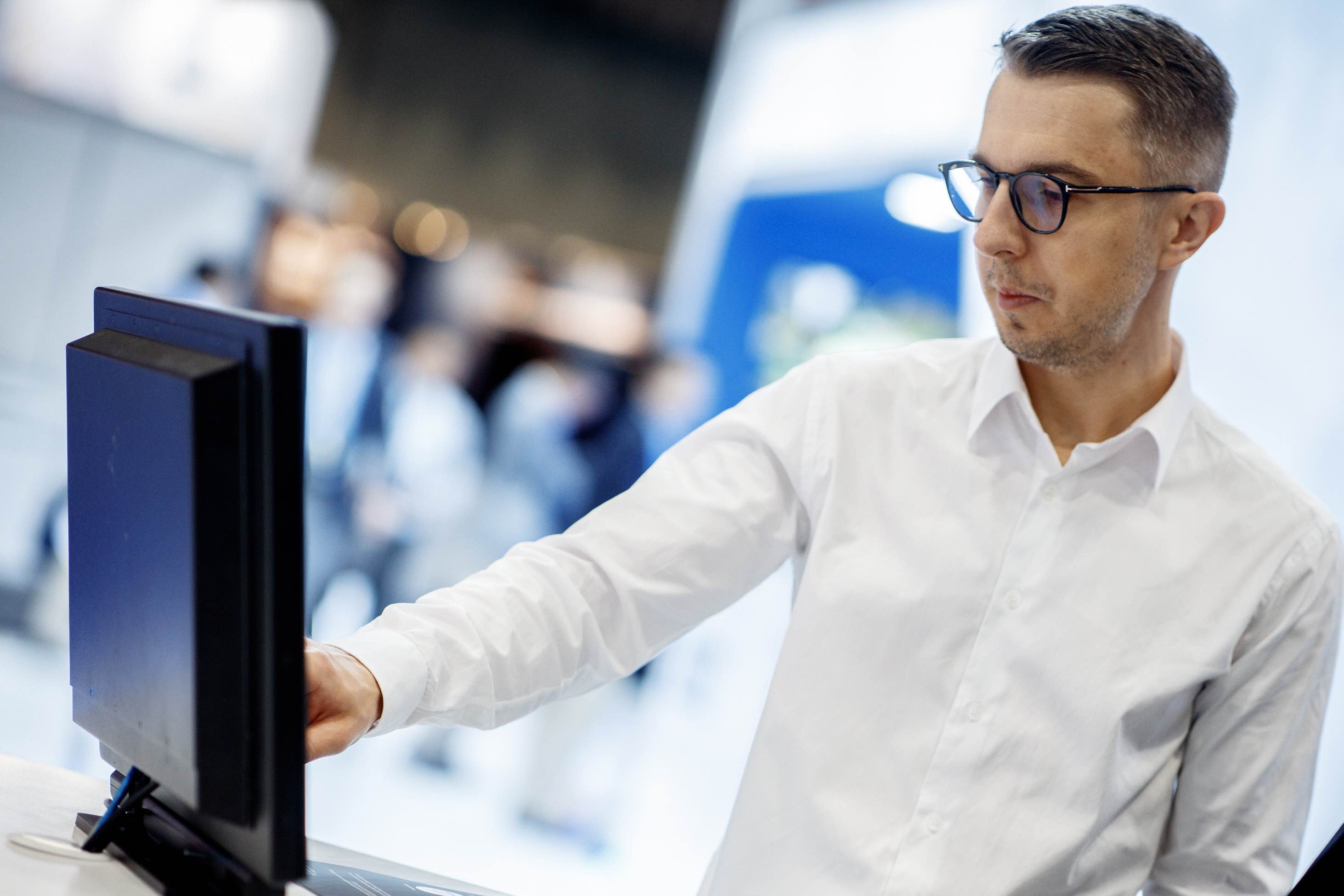 A male in a white shirt interacting with the touchscreen of an industrial Panel PC. We see the back of the Panel PC, at an angle. 