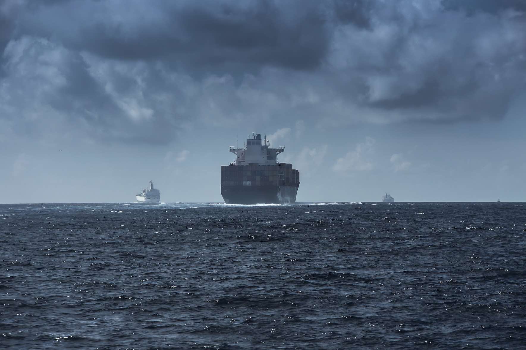 We see the stern of a cargo ship as it heads out the ocean, the weather looks rough. The picture has a blueish tint. 