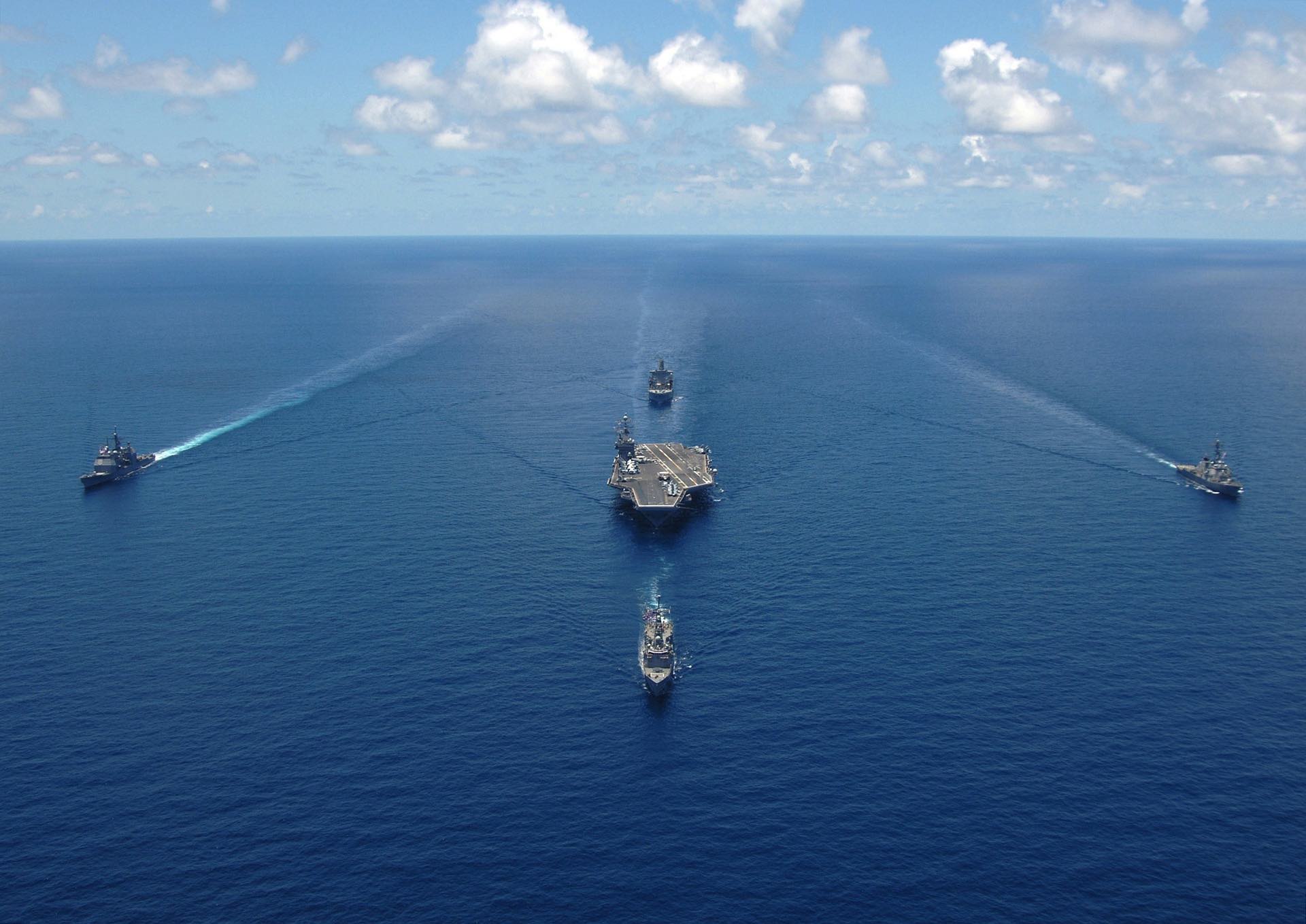 A naval fleet in formation, viewed from the sky. In the center of the picture is an aircraft carrier. It is being escorted by four other naval vessels. Blue sky, calm waters. 